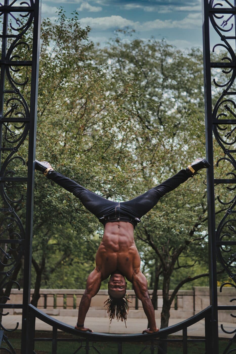 Alseny Sylla holding a freestanding handstand beneath an ornate iron pergola surrounded by trees