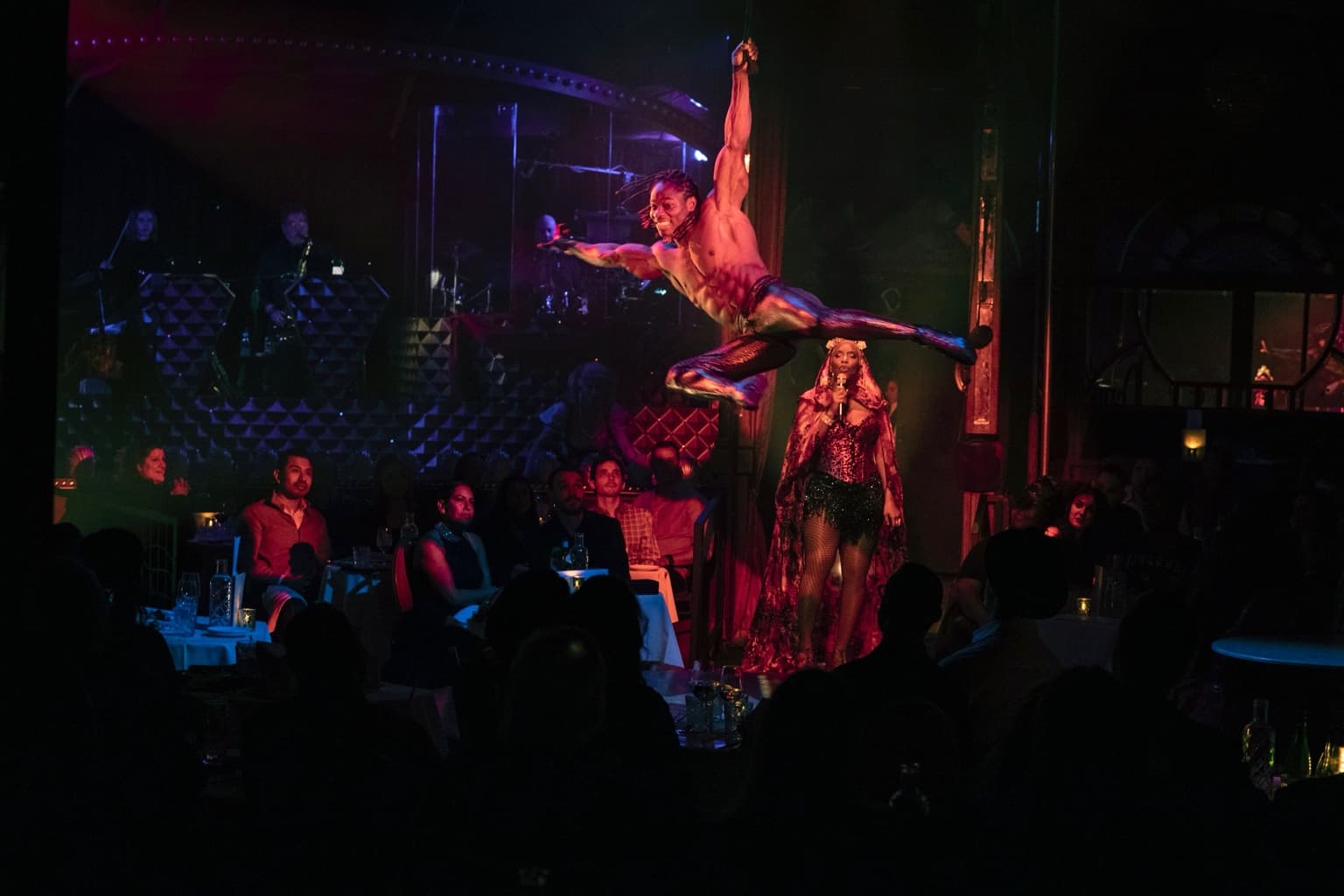 Alseny Sylla performing a one-arm aerial hold in a mid-air split above a live theatre audience